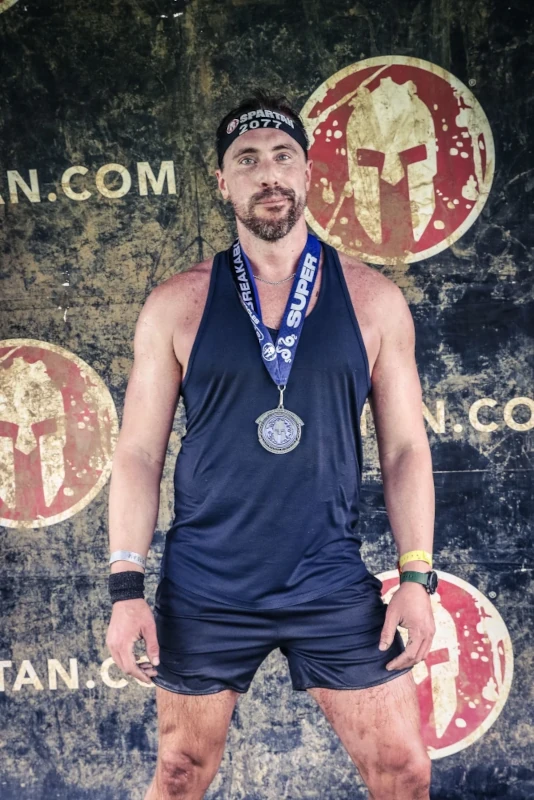 A muddy man with a medal in front of a spartan.com logo wall